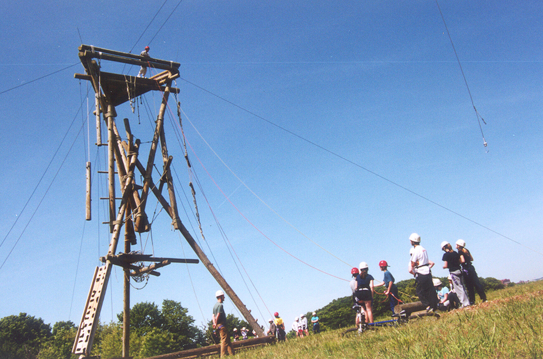 The image shows a tall wooden structure, likely part of an adventure course, with ropes and platforms. Several people wearing helmets are standing on a grassy hill, possibly waiting their turn. The sky is clear and blue, suggesting a sunny day. The overall scene conveys a sense of outdoor activity and adventure.